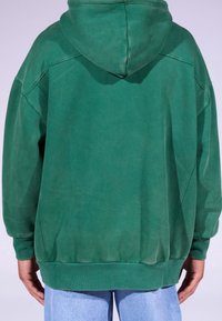 Felpa verde con cappuccio, in materiale misto cotone, vestibilità ampia, cappuccio grande, polsini e orlo a costine, caratterizzata da una superficie strutturata con variazioni distinte di colore.