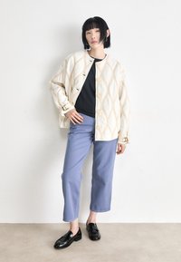 Colorful quilted jacket in cream with beige diamond pattern, paired with light blue trousers and black loafers. Buttons down the front.