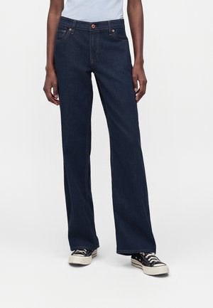 Άτομο που φοράει σκούρο μπλε τζιν με ίσια μπατζάκια, λευκό τοπ και μαύρα-άσπρα αθλητικά παπούτσια, στέκεται μπροστά σε απλό ανοιχτό φόντο.