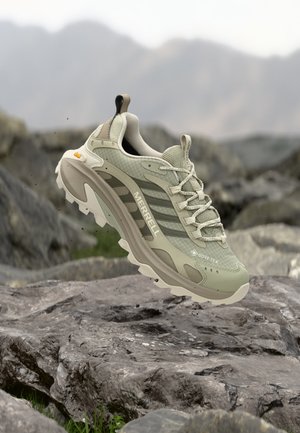 Hellgrüner Merrell-Wanderschuh mit Gore-Tex und Vibram-Sohle, der über felsigem Terrain in einer Outdoor-Bergumgebung schwebt.
