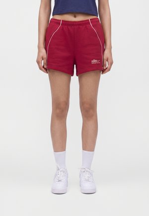 Personne portant un short de sport rouge Nike avec des liserés blancs, des chaussettes blanches et des baskets blanches, debout devant un fond uni.