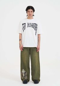 Witte oversized t-shirt met zwarte grafische tekst, gecombineerd met losse olijfgroene broek met een geborduurde ontwerp. Zwarte schoenen.