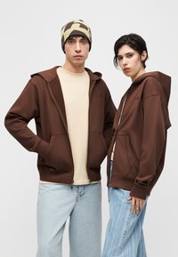 Δύο νέοι ενήλικες φορούν καφέ φούτερ με κουκούλα και ανοιχτόχρωμα μπλε τζιν, ο ένας με μπεζ σκουφάκι με σχέδια, στέκονται μπροστά σε ένα απλό φόντο.