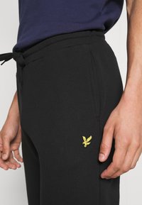 Schwarze Jogginghosen aus weichem, strukturiertem Stoff, mit einem elastischen Bund und einem Kordelzug sowie einem kleinen gelben gestickten Logo.