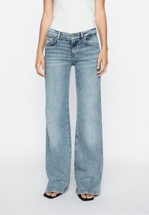 Femme portant un jean bleu clair à larges jambes et des sandales noires à bout ouvert, debout devant un fond blanc uni.