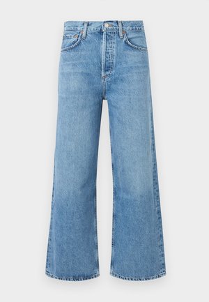 Frau steht in ärmellosem gestreiftem Rollkragenpullover, hoch taillierter weiter blauer Jeans mit schwarzem Gürtel und schwarzen offenen Schuhen vor einfarbigem Hintergrund.