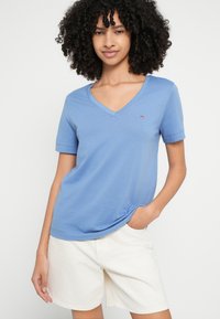 Jasnoniebieska koszulka z dekoltem w serek, wykonana z miękkiego materiału, z krótkimi rękawami, z małym haftowanym logo na lewej piersi. Noszona z białymi szortami.