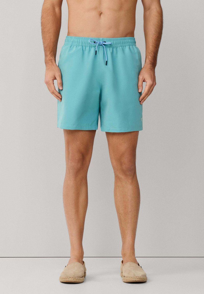 Homme portant un short de bain turquoise avec cordon de serrage et des chaussures beige à enfiler, debout devant un fond gris uni.