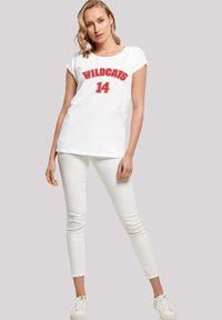 Balta medvilninė marškinė su raudonu užrašu "WILDCATS 14", trumpomis rankovėmis, derinama su baltais siaurais džinsais ir baltais sportbačiais.