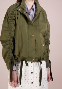 Olivgrön kort jacka med rynkade sid sömmar, guldknappar och justerbara band vid fållen och ärmarna. Har en hög krage.