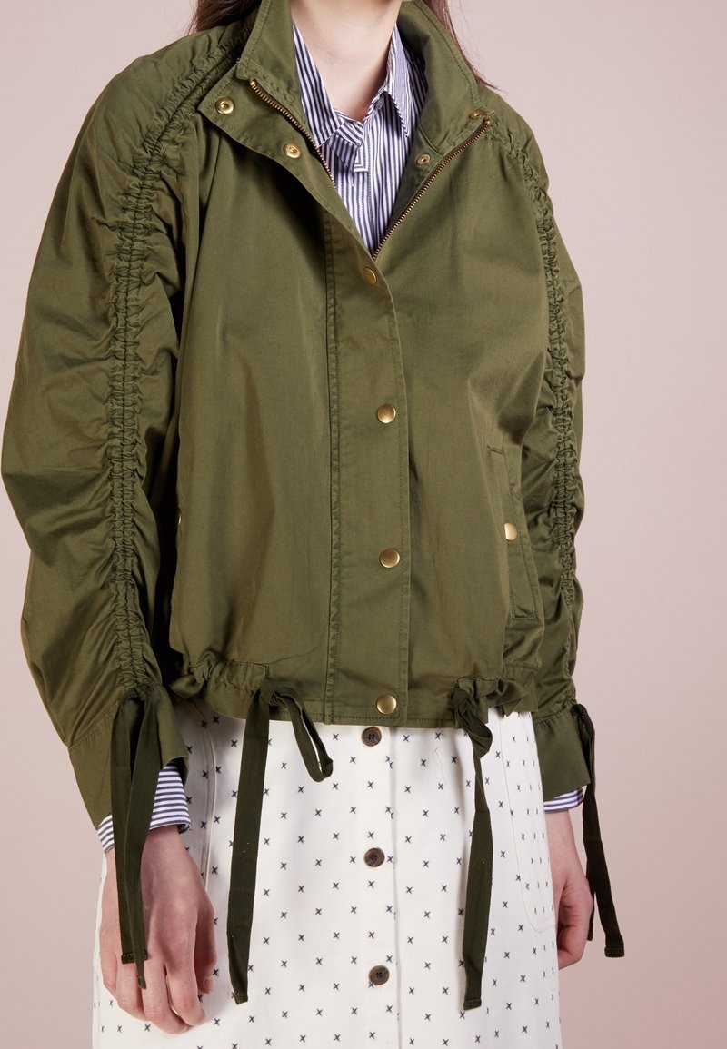 Olivgrön kort jacka med rynkade sid sömmar, guldknappar och justerbara band vid fållen och ärmarna. Har en hög krage.