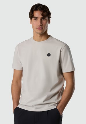 Giovane uomo con capelli castani corti e ondulati, che indossa una t-shirt beige chiaro a maniche corte con un piccolo logo circolare nero sul petto, con le mani in tasca.