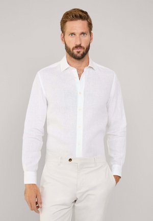 Chemise blanche en lin à manches longues avec col boutonné, présentant une patte de boutonnage à l'avant et un tissu texturé. Associée à un pantalon clair.