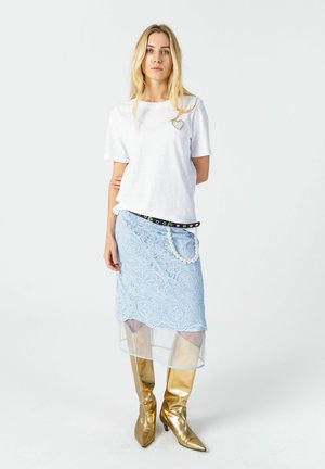 Vrouw die een wit T-shirt met korte mouwen en hertdetail draagt, lichtblauwe kanten rok met een transparante zoom, zwarte riem met parelketting en gouden kniehoge laarzen.