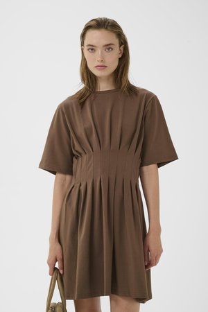 Kobieta z prostymi włosami nosi brązową sukienkę z krótkim rękawem i plisami w talii, trzymając beżową torbę na jednolitym tle.