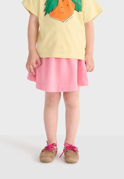 Enfant portant un t-shirt jaune avec un motif orange, une jupe rose et des chaussures marron avec des lacets rose vif, debout devant un fond uni.