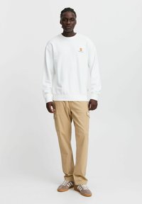 Felpa bianca con un piccolo logo arancione sul petto, abbinata a pantaloni cargo beige chiaro con tasche laterali e tessuto testurizzato.