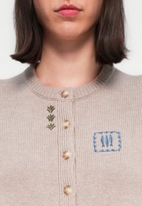 Beige gebreide cardigan met een ribbelstructuur, voorzien van geborduurde groene en blauwe details, en vijf ronde knopen aan de voorkant.