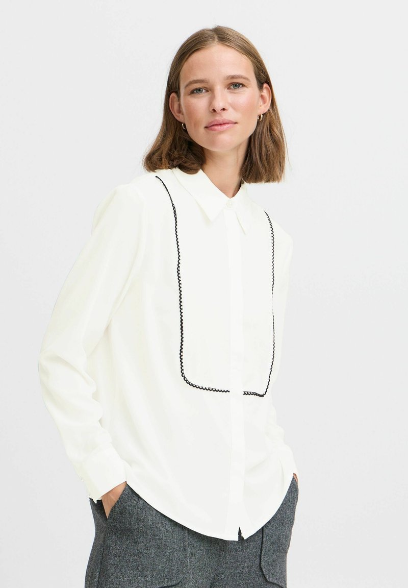 Camisa blanca de manga larga con un contorno rectangular negro bordado en el frente, con cuello puntiagudo y cierre de botones.