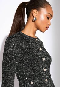 Svart paljettcardigan med rund halsringning, prydd med dekorativa knappdetaljer och en figurnära silhuett. Den glänsande texturen tillför visuell intresset.