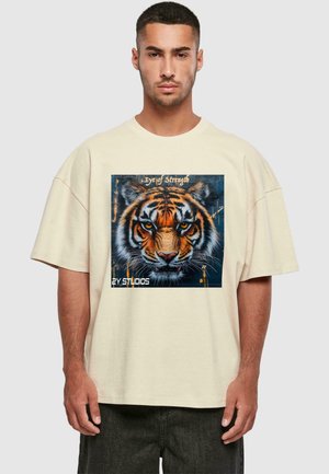 Beige oversized t-shirt featuring a vibrant tiger graphic with orange and black stripes against a dark background, and text "Eye of Strength."