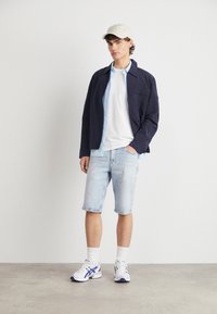 Giacca leggera della marina sopra una camicia azzurra, t-shirt bianca, pantaloni corti di denim chiaro, calze bianche e scarpe da ginnastica. Sfondo chiaro.