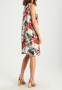 Vestido de flores sin mangas en blanco con patrones tropicales en rosa y verde. Corte holgado con un detalle de flecos en el dobladillo. La modelo lleva sandalias metálicas de tacón cuadrado.