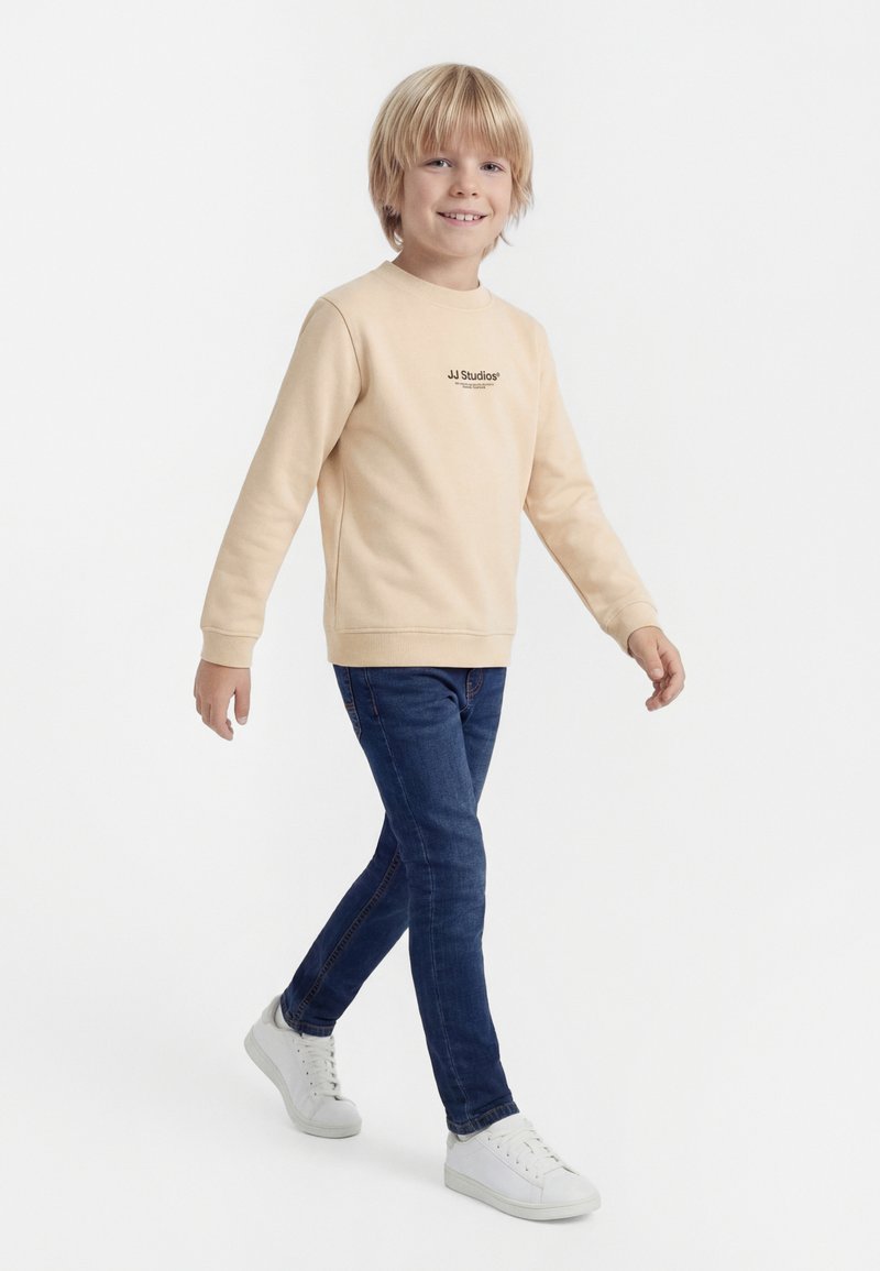 Ragazzo con capelli biondi che cammina, indossa una felpa beige, jeans blu scuro e scarpe da ginnastica bianche su uno sfondo bianco semplice.