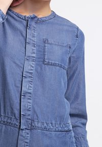 Jeansskjorta i ljusblått, med rund hals, knappstängning framtill, en bröstficka och elastisk midja, med långa ärmar.