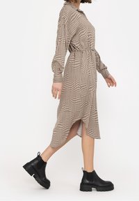 Brown and beige checkered dress with long sleeves, collared neck, belted waist, and asymmetric hem. Paired with chunky black ankle boots.
