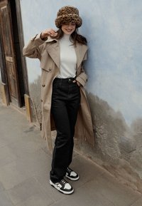 Donna in trench beige, dolcevita bianco, jeans neri e cappello a stampa leopardata sorride mentre sistema il cappello vicino a una parete texturizzata blu e grigia.