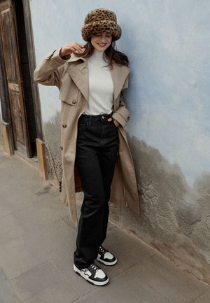 Mujer con abrigo trinchera beige, cuello alto blanco, jeans negros y sombrero de estampado de leopardo sonríe mientras ajusta su sombrero cerca de una pared texturizada azul y gris.