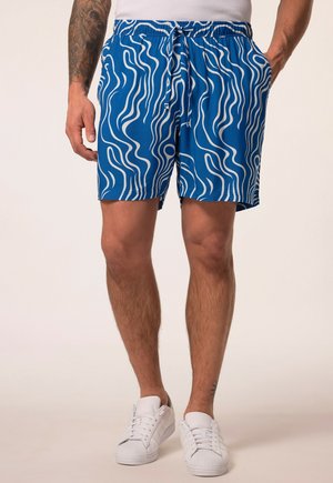 Mand iført blå badeshorts med hvide bølgede linjer, hvide sneakers og en hvid tanktop, står med den ene hånd i lommen.