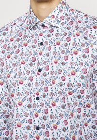 Chemise blanche à boutons avec un motif floral coloré comprenant des fleurs roses, jaunes et bleues. Tissu lisse avec un col standard.