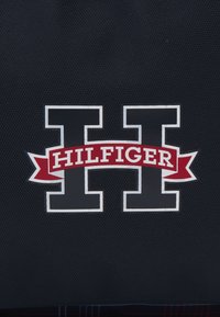 Il tessuto texturato blu navy presenta una grande lettera "H" in grigio scuro, con un nastro rosso sottostante che riporta la parola "HILFIGER" in bianco.