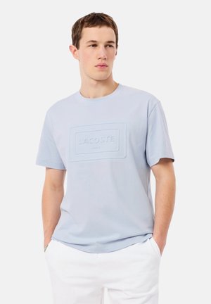 Junger Mann trägt ein hellblaues T-Shirt mit geprägtem Lacoste-Logo und weiße Hose, steht mit den Händen in den Taschen und schaut seitlich weg.