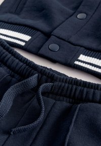 Temno modra bombažna jopa za tracksuit z ježki na prsih in črtastimi rebrastimi manšetami, združena z ujemajočimi se hlačami z vezalko in rebrastim pasom.