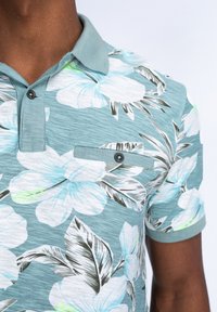 T-paita lyhyillä hihoilla turkoosina kukkakuviolla, jossa on suuria valkoisia ja sinisiä kukkia, rintatasku ja tummat napit.