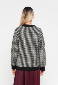 Woman with long ombre hair wearing a black and white houndstooth sweater and a burgundy skirt, facing away against a white background.