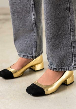 Zapatos de tacón bajo en dorado y negro con un acabado metálico y puntera de ante negro, combinados con jeans de mezclilla gris a la altura del tobillo.