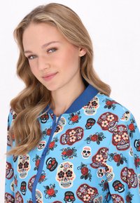 Jachetă albastră cu modele ce includ craniene colorate și motive florale, cu guler ribbat și închidere cu fermoar în față. Textură de țesătură moale.