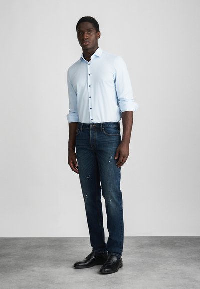 Ανοιχτό μπλε πουκάμισο με κουμπιά, σκούρες μπλε τζιν, μαύρα παπούτσια. Το πουκάμισο έχει μακριά μανίκια και απλό μανίκι, τα τζιν διαθέτουν φθαρμένα στοιχεία.