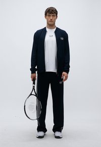 Completo sportivo blu navy con giacca con cerniera e pantaloni abbinati, abbinato a una maglietta bianca. Modello che tiene in mano una racchetta da tennis.