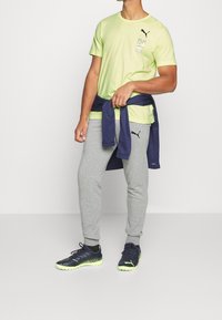 Camiseta amarilla de Puma con logo, pantalones de chándal grises, sudadera azul oscura atada a la cintura y zapatillas deportivas negras con acentos neón.