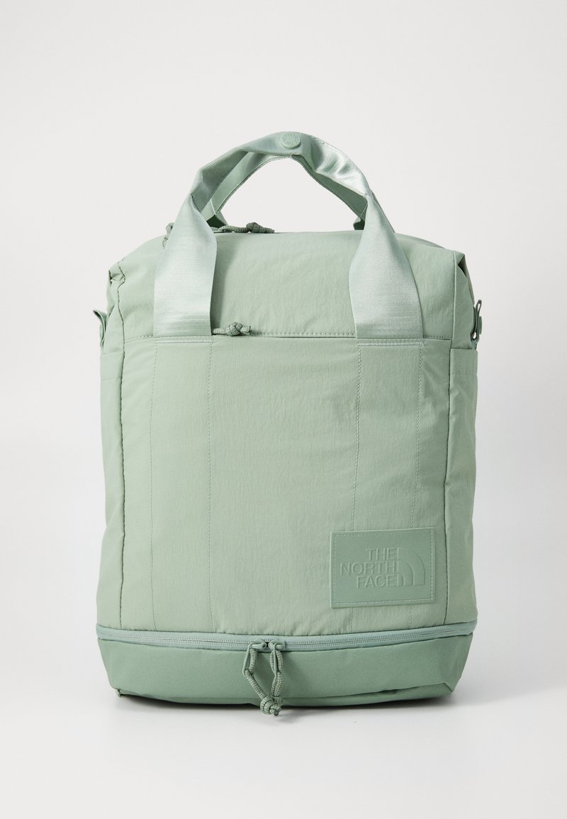 Mochila de tela verde con dos asas superiores, cierre de cremallera y un parche de logotipo en la parte frontal. Presenta una textura suave y un cordón ajustable en la parte inferior.