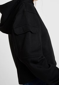 Czarna, skrócona bluza z kapturem wykonana z miękkiego materiału, z dużym kapturem i kieszenią na lewym rękawie. Gładka faktura, minimalystyczny design.