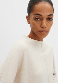 Frau mit zurückgebundenem dunklem Haar, die einen cremefarbenen Pullover trägt, der vorne eine sichtbare Naht aufweist, vor einem schlichten hellen Hintergrund.