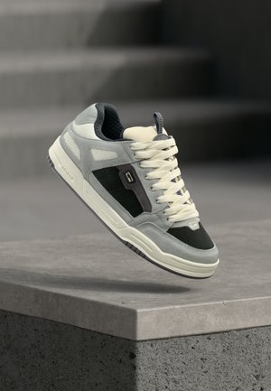 Grauer und schwarzer Turnschuh mit weißen Schnürsenkeln, der über einer Betonbrüstung schwebt, verschwommene Treppen im Hintergrund.