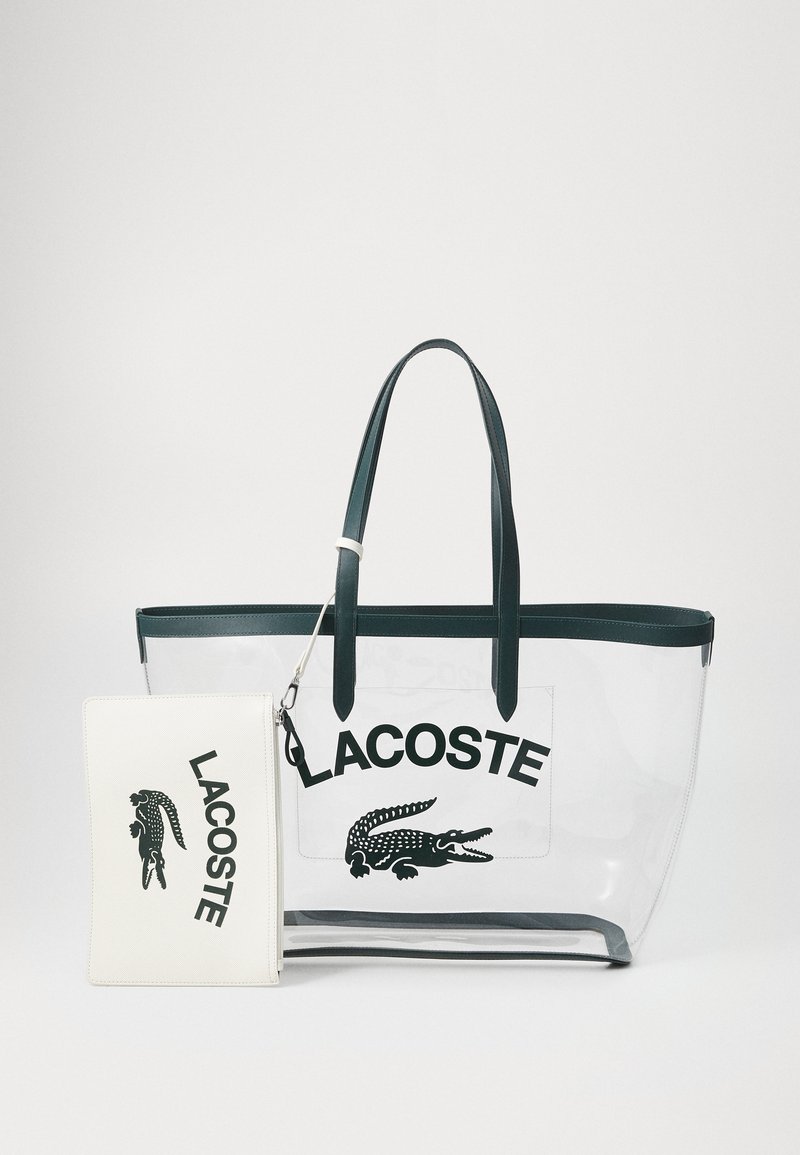 Clear tote bag with dark green handles, featuring a white zip pouch. Black brand logo and crocodile graphic prominently displayed.
