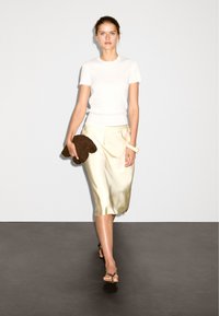 Bílý přiléhavý tričko, saténová krémová midi sukně, hnědá texturovaná kabelka a sandály na pásky. Minimalistický design s jemnými texturami a barvami.